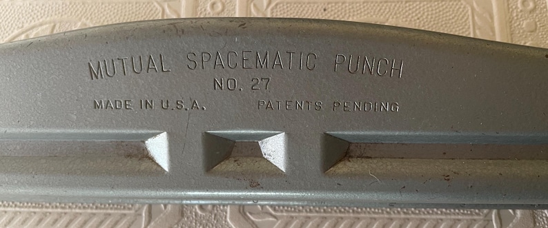May include: Close-up of a vintage gray metal paper punch. The text "MUTUAL SPACEMATIC PUNCH" and "NO. 27" are visible. The words "MADE IN U.S.A." and "PATENTS PENDING" are also present. The punch has a curved top and three rectangular punch holes.