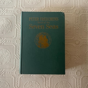 May include: A hardcover book titled "Peter Freuchen's Book of the Seven Seas" in dark green with gold lettering and a gold emblem. The book is closed and appears to be in good condition.