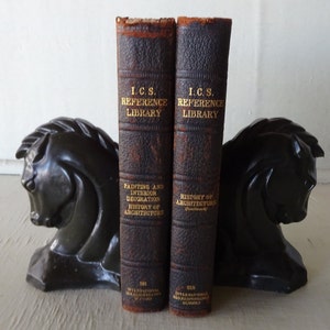 May include: Two vintage brown leather-bound books with gold lettering titled "I.C.S. Reference Library" and "Painting and Interior Decoration History of Architecture" and "History of Architecture"  are being held up by a pair of black horse bookends.