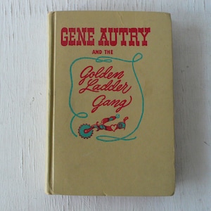 May include: A vintage paperback book with a yellow cover featuring the text "GENE AUTRY AND THE Golden Ladder Gang" in red and blue lettering. The book cover also features a stylized illustration of a wagon wheel and a heart.