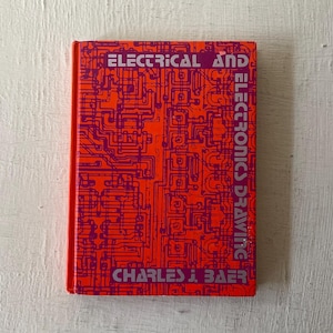 May include: A hardcover book titled "ELECTRICAL AND ELECTRONICS DRAWING" by Charles J. Baer. The cover is a vibrant orange with a purple circuit board design. The book is rectangular and has a red spine.