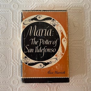 May include: A vintage book titled "Maria: The Potter of San Ildefonso" by Alice Marriott. The book has an orange and black cover with a circular design featuring stylized bird and geometric shapes. The title is in elegant script.