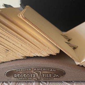 May include: A vintage Rolodex file with a metal base and a fan of beige index cards. The Rolodex has the text "ZEPHYR AMERICAN ROLODEX V-FILE Jr." on the front. The cards are held in place by metal clips.