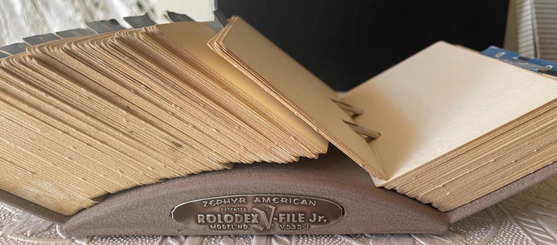 May include: A vintage Rolodex file with a metal base and a fan of beige index cards. The Rolodex has the text "ZEPHYR AMERICAN ROLODEX V-FILE Jr." on the front. The cards are held in place by metal clips.