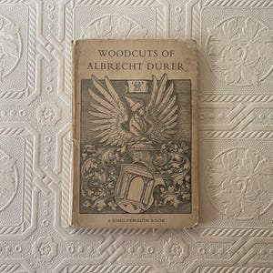 May include: A vintage book titled "Woodcuts of Albrecht Dürer" with a detailed illustration on the cover. The cover art features a winged figure and a coat of arms. The book is a King Penguin Book.