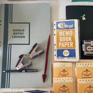 May include: An assortment of vintage office supplies, including a ledger, memo book paper, paper clips, and record forms. The ledger is light blue with the words "SINGLE ENTRY LEDGER" in a black frame. Other items include staplers, a pen, and a red arrow-shaped paper clip.