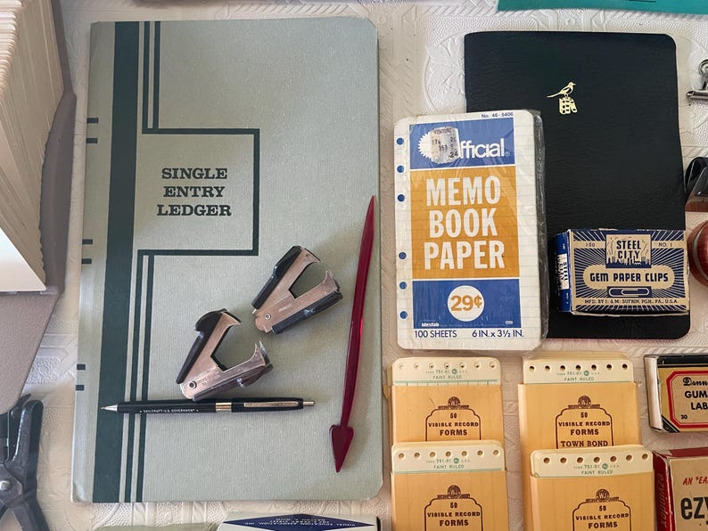 May include: An assortment of vintage office supplies, including a ledger, memo book paper, paper clips, and record forms. The ledger is light blue with the words "SINGLE ENTRY LEDGER" in a black frame. Other items include staplers, a pen, and a red arrow-shaped paper clip.