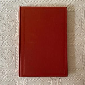 May include: A hardcover book with a solid red cover. The cover has a rectangular border and a slightly textured appearance. The book is lying on a white patterned surface.
