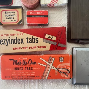May include: A collection of vintage office supplies, including boxes of gummed labels, staples, and index tabs. The boxes are in various colors, with text labels. A dark brown Bates index card file is also visible.
