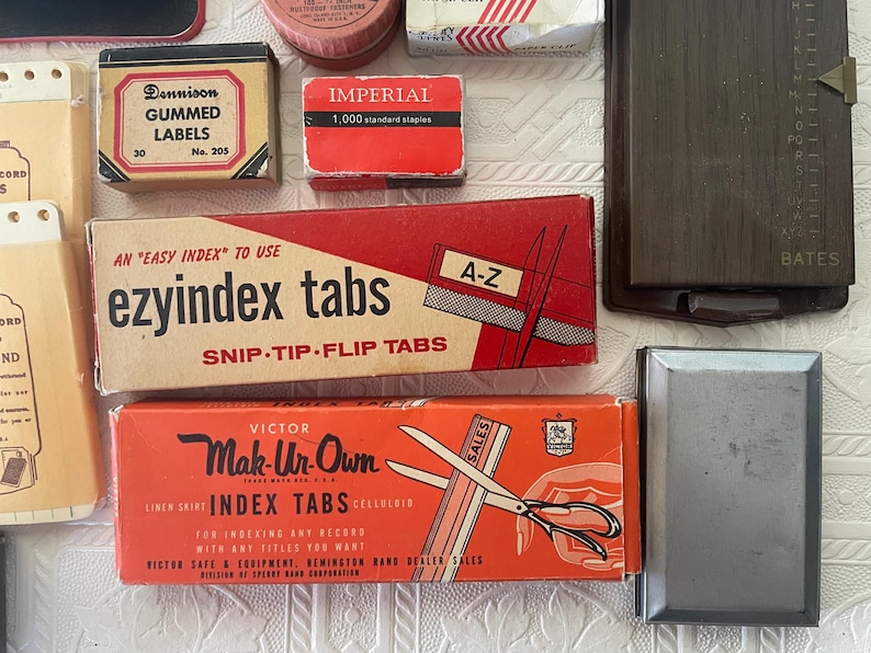 May include: A collection of vintage office supplies, including boxes of gummed labels, staples, and index tabs. The boxes are in various colors, with text labels. A dark brown Bates index card file is also visible.
