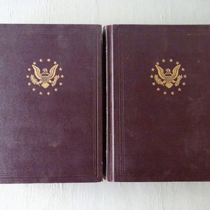 May include: Two dark brown hardcover books with gold embossed designs on the front covers. Each cover features a gold eagle with a shield and a wreath of leaves, surrounded by a circle of gold stars.