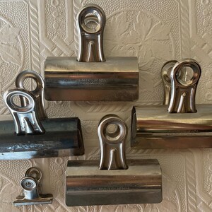 May include: A collection of silver metal binder clips of varying sizes. The clips have a rounded handle and a spring-loaded mechanism. Some clips have the text "HUNT CLIP CO." imprinted on them. The clips are arranged on a textured, off-white surface.