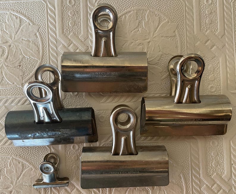 May include: A collection of silver metal binder clips of varying sizes. The clips have a rounded handle and a spring-loaded mechanism. Some clips have the text "HUNT CLIP CO." imprinted on them. The clips are arranged on a textured, off-white surface.