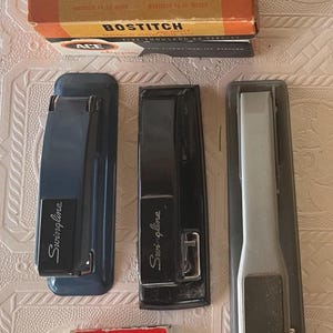 May include: An assortment of vintage office supplies, including three staplers in blue, black, and gray, and a box of Bostitch staples. A small box of Imperial staples is also present.