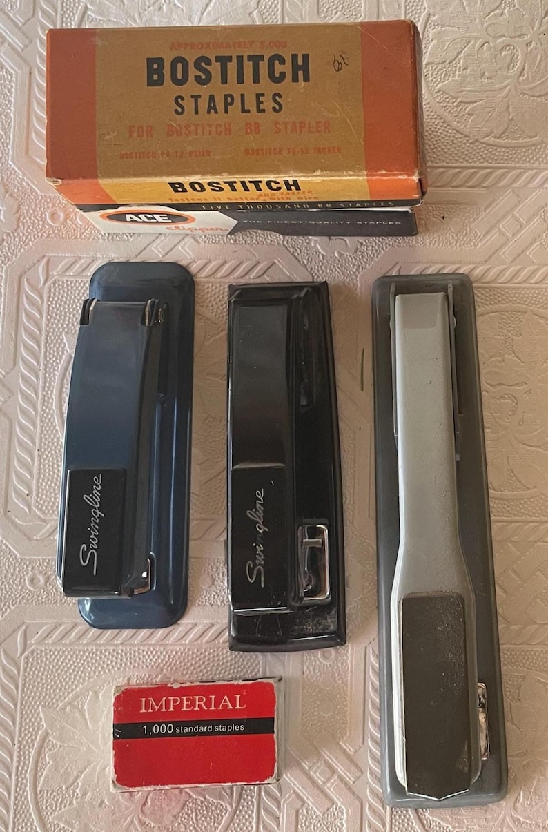May include: An assortment of vintage office supplies, including three staplers in blue, black, and gray, and a box of Bostitch staples. A small box of Imperial staples is also present.