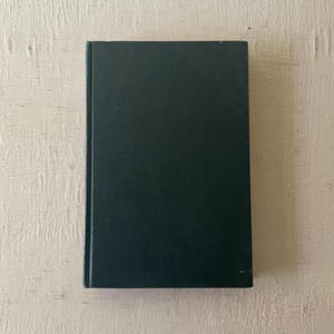 May include: A dark green hardcover book with a textured cover. The book is rectangular and appears to be closed, with the spine visible on the left side. The background is a light-colored, textured surface.
