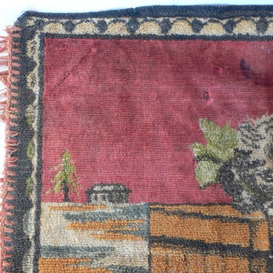 May include: A close-up of a red, brown, and green tapestry with a woven design of a house, trees, and a floral motif. The tapestry has a black and white fringe border.