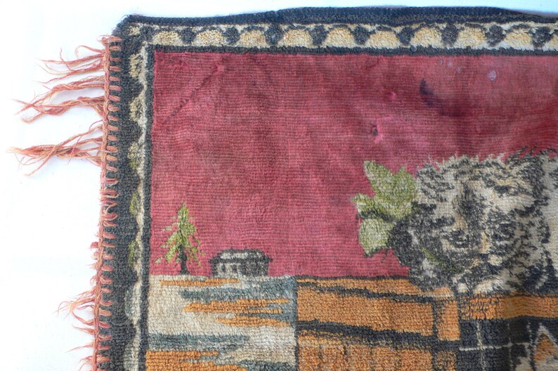 May include: A close-up of a red, brown, and green tapestry with a woven design of a house, trees, and a floral motif. The tapestry has a black and white fringe border.