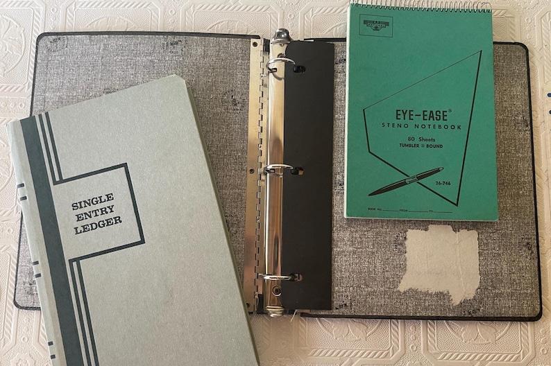 May include: An open gray binder with a silver metal spine, a light blue ledger with the text "SINGLE ENTRY LEDGER", and a green "EYE-EASE" steno notebook with a pen illustration. The binder and notebooks are on a patterned surface.