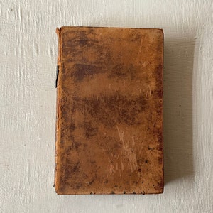 May include: A vintage book with a worn, brown leather cover. The cover shows signs of age with dark spots and a distressed texture. The book's spine is visible, and the edges are slightly frayed, suggesting it is an antique.