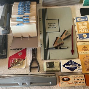 May include: An assortment of vintage office supplies, including staplers, a hole punch, index card files, a ledger, and boxes of staples. The items are in various colors, such as black, gray, and blue, and are arranged on a white surface.