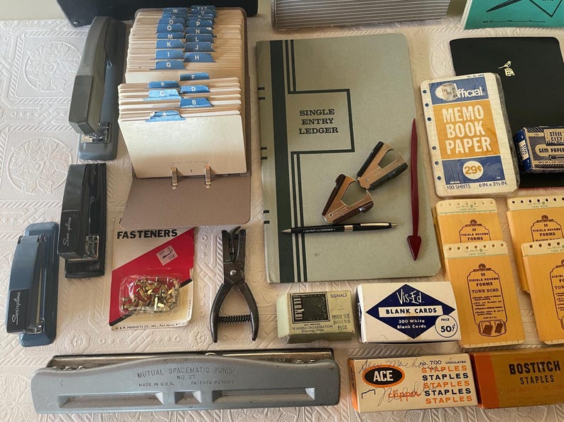 May include: An assortment of vintage office supplies, including staplers, a hole punch, index card files, a ledger, and boxes of staples. The items are in various colors, such as black, gray, and blue, and are arranged on a white surface.