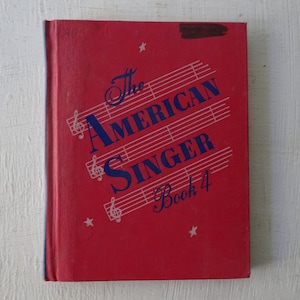 May include: The cover of a red book titled "The American Singer Book 4". The title is in blue lettering with musical notes and stars printed on the cover.