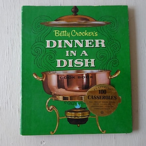 May include: A vintage cookbook titled "Betty Crocker's Dinner in a Dish" with a green cover. The cover features a copper-colored chafing dish illustration and the text "Featuring 100 Casseroles".