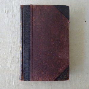 May include: A vintage, brown leather-bound book with a black spine and gold lettering. The book is closed and sitting on a white surface.