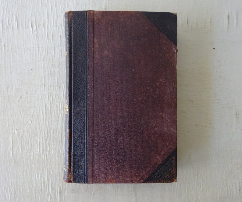 May include: A vintage, brown leather-bound book with a black spine and gold lettering. The book is closed and sitting on a white surface.