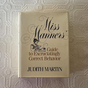 May include: A hardcover book titled "Miss Manners" with a cream-colored cover. The title is in elegant script above the subtitle "Guide to Excruciatingly Correct Behavior." The author's name, Judith Martin, is at the bottom. An illustration of a woman at a typewriter is also on the cover.