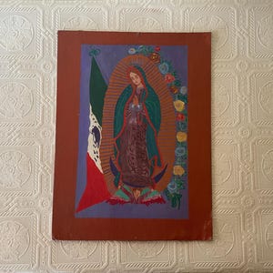 May include: A rectangular painting on a dark red background featuring the Virgin of Guadalupe in a green cloak and brown dress, standing on a crescent moon held by an angel. To the left is a Mexican flag, and to the right is a floral garland.