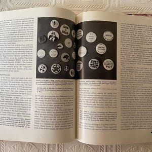 Puede incluir: Libro abierto con una p&aacute;gina doble que muestra una colecci&oacute;n de botones blancos con texto y gr&aacute;ficos en negro. Los botones tienen frases como "God is Dead" y "Legalize Pot". Los n&uacute;meros de p&aacute;gina son 66 y 67.