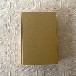May include: A hardcover book with a tan cover. The book is rectangular and has a smooth, solid color. The background is a white, textured surface with a repeating floral pattern. The book is centered in the image.