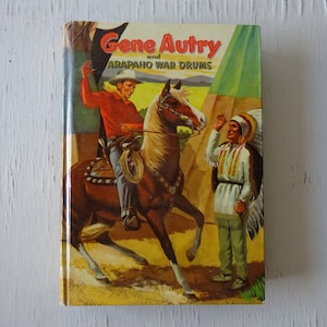 May include: A vintage book cover featuring Gene Autry on horseback, wearing a cowboy hat and red shirt, with a Native American man in traditional clothing standing to the right. The book title is "Gene Autry and Arapaho War Drums".