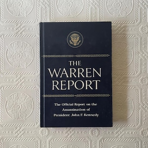 May include: A hardcover book titled "The Warren Report" with the subtitle "The Official Report on the Assassination of President John F. Kennedy." The cover is navy blue with gold text and a gold presidential seal.