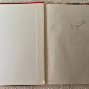 Puede incluir: Libro abierto con cubierta roja y p&aacute;ginas blancas. La p&aacute;gina derecha tiene una firma manuscrita en tinta azul. El libro est&aacute; abierto sobre una superficie estampada.