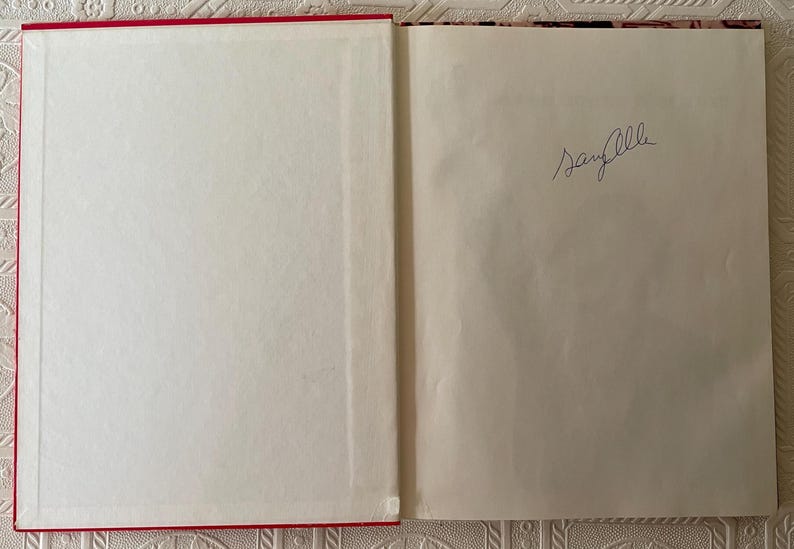 Puede incluir: Libro abierto con cubierta roja y p&aacute;ginas blancas. La p&aacute;gina derecha tiene una firma manuscrita en tinta azul. El libro est&aacute; abierto sobre una superficie estampada.