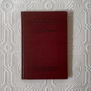 May include: A maroon-colored book titled "A Short History of Navigation" with gold lettering. The book is closed and has a textured cover. The book is on a white patterned surface.