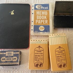 May include: A collection of vintage office supplies, including a black notebook with a gold bird emblem, memo book paper, a black ink pad, and boxes of paper clips and record forms. The memo book paper is 6 x 3.5 inches.