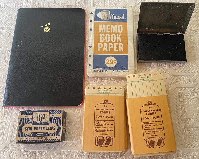 May include: A collection of vintage office supplies, including a black notebook with a gold bird emblem, memo book paper, a black ink pad, and boxes of paper clips and record forms. The memo book paper is 6 x 3.5 inches.