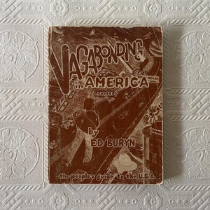 May include: A vintage book titled "Vagabonding in America" by Ed Buryn. The cover is brown with white lettering and illustrations of a person with a backpack, cars, and buildings. The subtitle reads "the people's guide to the U.S.A."