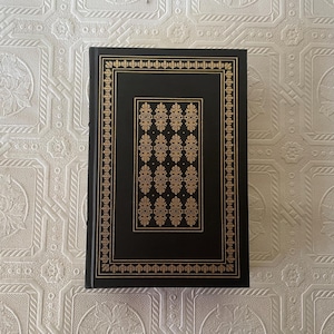 May include: A hardcover book with a black cover and gold decorative accents. The cover features a gold border and a central rectangular design with repeating gold floral patterns. The book is on a white patterned surface.