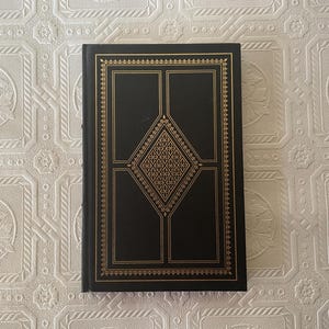 May include: A hardcover book with a black cover and gold decorative accents. The cover features a geometric design with a diamond pattern in the center and a gold border. The book is closed and appears to be in good condition.