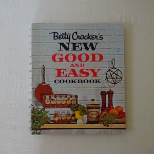 May include: A vintage cookbook titled "Betty Crocker's New Good and Easy Cookbook" with a brick background. The cover features a variety of kitchen items, including a copper pot, spice jars, a cookie jar, and a metal rack.