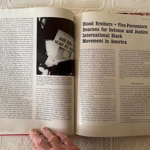 Puede incluir: Un libro abierto con tapas rojas, que muestra texto y una fotograf&iacute;a en blanco y negro. El texto incluye el t&iacute;tulo "Blood Brothers &bull; Five-Percenters Deacons for Defense and Justice International Black Movement in America."