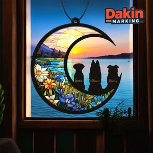 May include: A black crescent moon-shaped ornament with a stained-glass design. The ornament features silhouettes of three dogs, the names Guwi, Alain, and Henry, and a sunset scene. The text "Dakin Marking" is visible.