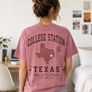 Puede incluir: Una camiseta de manga corta color rosa polvoriento con un gráfico en la espalda que incluye el texto COLLEGE STATION, un mapa de Texas con una estrella, EST. 1938 y el texto TEXAS Home of Big Dreams en letras granates con estrellas.