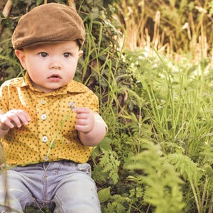 May include: A young child wearing a brown corduroy cap, a yellow button-down shirt with black polka dots, and blue jeans sits in tall green grass. The child is holding a small flower.