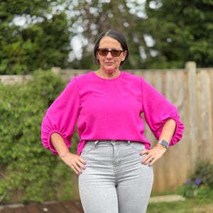 May include: A woman wearing a bright pink blouse with long, gathered sleeves and light gray jeans. The blouse has a round neckline. The woman is wearing sunglasses and a watch. The background includes green foliage and a wooden fence.